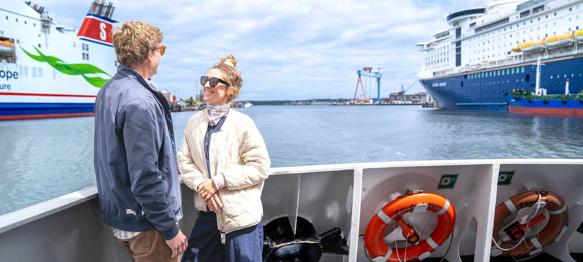 Zwei Personen auf einem Schiff mit der Förde und zwei großen Fähren im Hintergrund