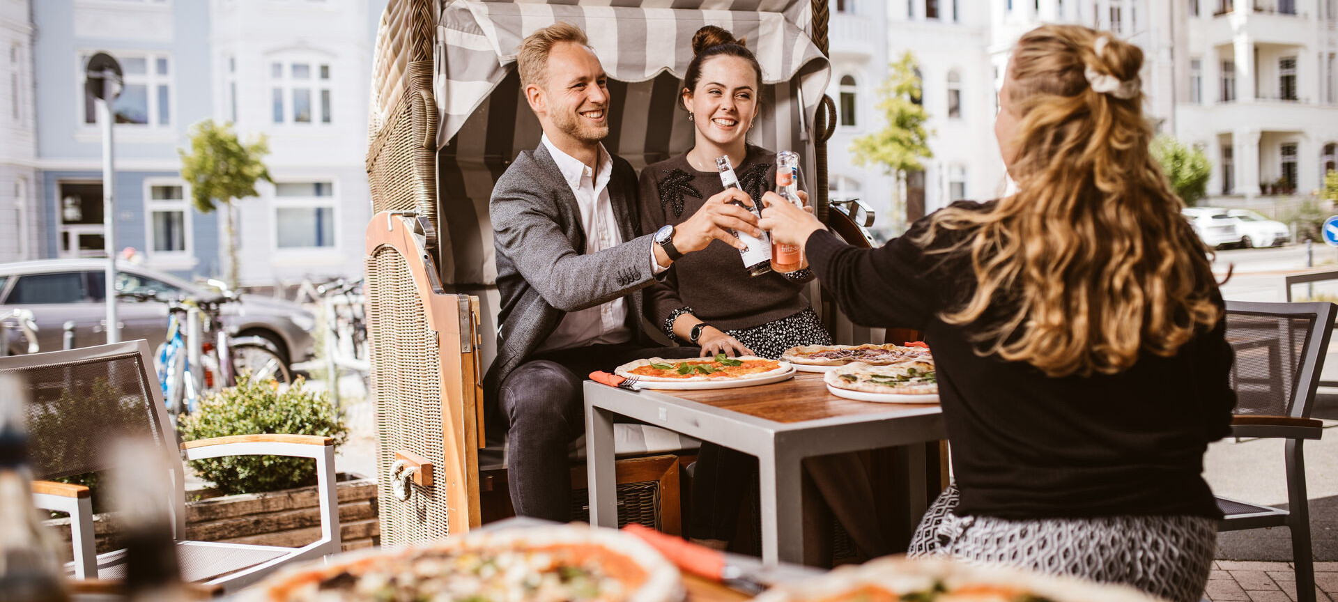 Drei Freund:innen essen zusammen Pizza und stoßen mit ihren Getränken an. 