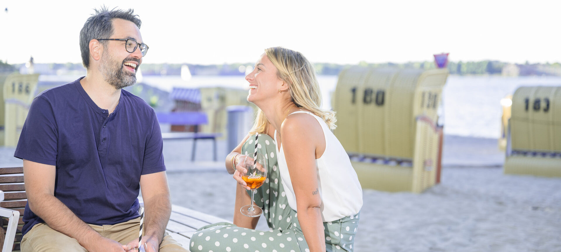 Pärchen sitzt lächelnd am Strand in Kiel mit Cocktails in der Hand, im Hintergrund sind viele Strandkörbe