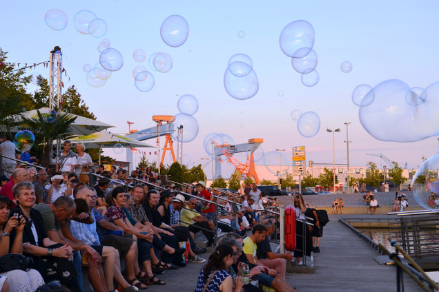 Das Publikum sitzt am frühen Abend auf Stufen am Bootshafen, große Seifenblasen in der Luft